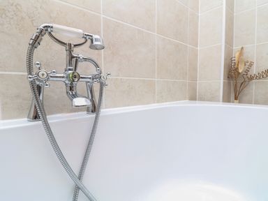A bathtub with a chrome faucet and handheld showerhead with beige tiled walls and a dried flower arrangement in the corner at The Croft in Holsworthy