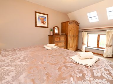 A bedroom with a bed covered in a patterned bedspread two folded towels wooden wardrobe chest of drawers with mirror window with curtains and skylights at The Croft in Holsworthy