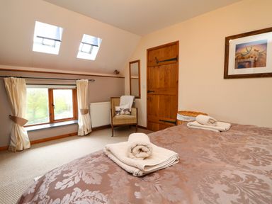 A bedroom with a bed covered in patterned bedding and folded towels a wooden door a chair and windows with curtains at The Croft in Holsworthy