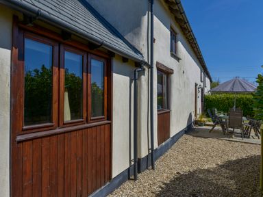 An exterior view of a building with wooden framed windows a gravel area and outdoor seating with an umbrella at The Croft in Holsworthy