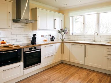 A kitchen with white cabinets wooden countertops a microwave oven a built in oven a stove a kettle a knife block a vase with flowers and a window at Holly Edge in Moorsholm