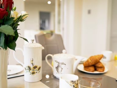A table set with a white teapot, a coffee pot with sunflower design, cups, a vase with red and white roses, and a plate of croissants at Holly Edge in Moorsholm