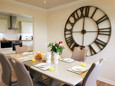 A dining area with a glass table set with plates and glasses near a kitchen and a large wall clock at Holly Edge in Moorsholm
