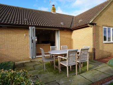 An outdoor patio with a wooden table and six chairs outside a brick house with open folding doors at Holly Edge in Moorsholm
