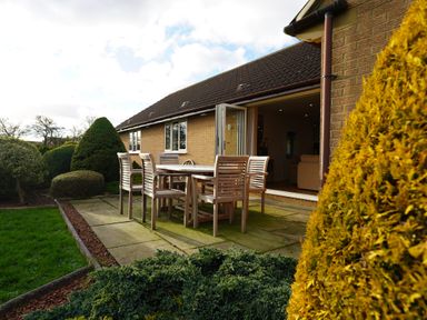 A patio with a wooden table and six chairs outside a house with an open door and garden shrubs at Holly Edge in Moorsholm