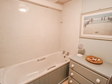 A bathroom with a bathtub tiled walls a cabinet with drawers a small round mirror and a framed beach picture at Janes Place in Lifton