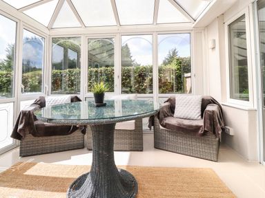 A conservatory with a glass round table and three wicker chairs with cushions at Palm Court in Burton-in-Kendal