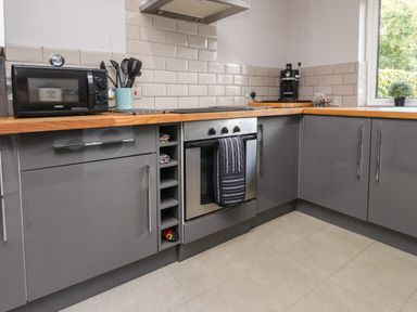 A kitchen with grey cabinets wooden countertops a microwave an oven cooking utensils a coffee machine and a window with a plant at Palm Court Burton-in-Kendal