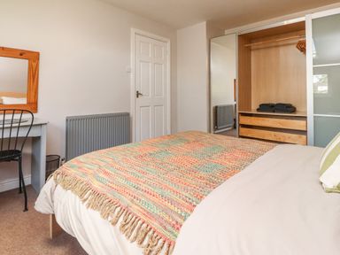 A bedroom with a bed covered by a multicolored blanket a desk with a mirror and chair an open wardrobe with shelves and hanging space at Palm Court in Burton-in-Kendal