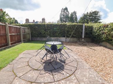 A backyard with a round metal table and chairs on a stone patio a grass area a pebble area with a rotary clothesline and wooden fencing and hedges