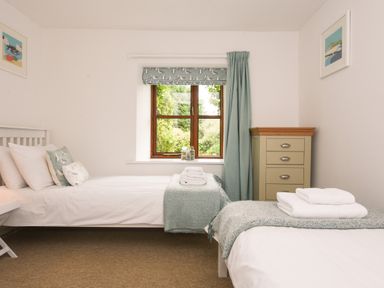 A bedroom with two single beds with white linens and teal blankets under a window with teal curtains and a beige chest of drawers at Lily Cottage in Looe