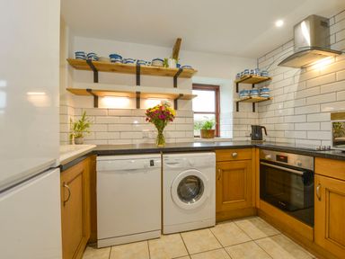 A kitchen with white appliances wooden cabinets open shelves with dishes and a bouquet of flowers on the counter at Lily Cottage in Looe