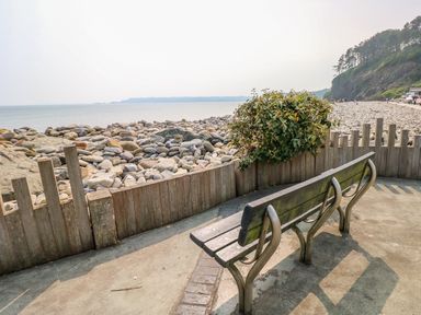A wooden bench on a paved area overlooking a rocky beach with a cliff and sea in the background at Swn y mor in Amroth