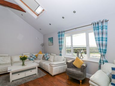A living room with white leather sofas a grey armchair a white coffee table with flowers and blue check curtains at Swn y mor in Amroth