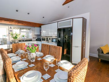 A dining area with a wooden table set with white dishes and glasses next to a kitchen with white cabinets and a black refrigerator at Swn y mor in Amroth