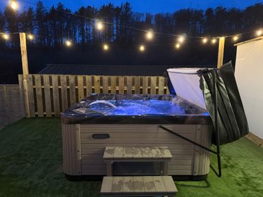 An outdoor hot tub with water jets and steps on artificial grass with string lights above at Swn y mor in Amroth