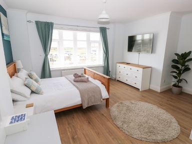 A bedroom with a wooden bed, pillows and towels, a dresser with a mounted TV, an oval rug, curtains, and a potted plant at Swn y mor in Amroth