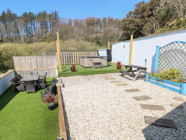 An outdoor patio area with a hot tub a picnic table wicker chairs around a glass table and a gravel walkway at Swn y mor in Amroth