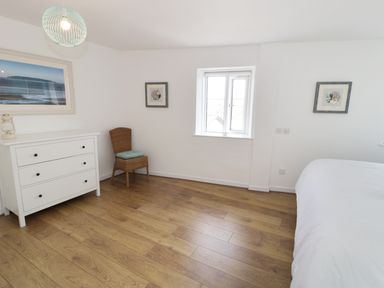 A bedroom with wooden floor a white dresser a wicker chair with a cushion wall art and a window at Waverley House in Beaumaris