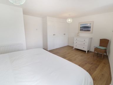 A bedroom with a white bed, white wardrobes, a white chest of drawers, a wicker chair with a cushion and a framed picture on the wall at Waverley House in Beaumaris