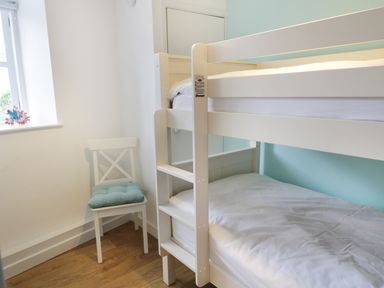 A small bedroom with white bunk beds a white chair with a cushion and a window at Waverley House in Beaumaris