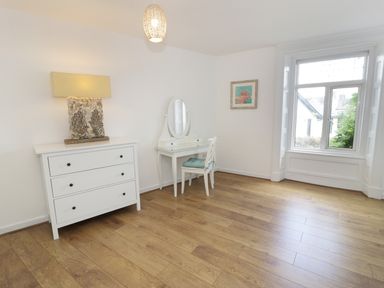 A room with wooden floor a white chest of drawers with lamp a white dressing table with mirror and chair and a window at Waverley House in Beaumaris
