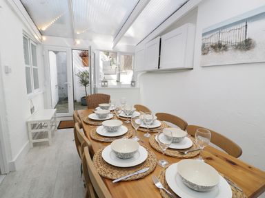 A dining area with a wooden table set with bowls and glasses and wicker chairs in a room with glass ceiling at Waverley House in Beaumaris