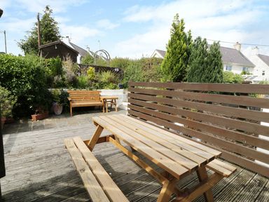 An outdoor wooden picnic table and bench with wooden bench seating and plants in the background at Waverley House in Beaumaris