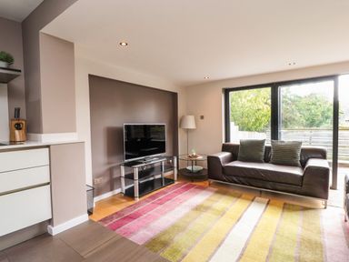 A living room with a brown leather sofa two cushions a TV on a stand a round side table and large sliding glass doors at Underfell in Burton-In-Kendal