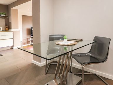 A dining area with a glass table metal legs and two translucent chairs a small plant on the table and a kitchen visible in the background at Underfell in Burton-In-Kendal