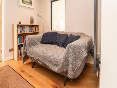 A small living area with a gray sofa covered by a gray throw with dark cushions a wooden bookshelf with books a floor lamp and a painting on the wall at Underfell in Burton-In-Kendal