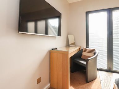A room corner with a wall-mounted TV a wooden desk with a mirror a black chair with a striped pillow and glass doors with blinds at Underfell in Burton-In-Kendal