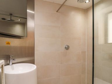 A bathroom with a circular white sink a faucet a mirror a showerhead control on beige tiled walls and a glass shower partition at Underfell in Burton-In-Kendal