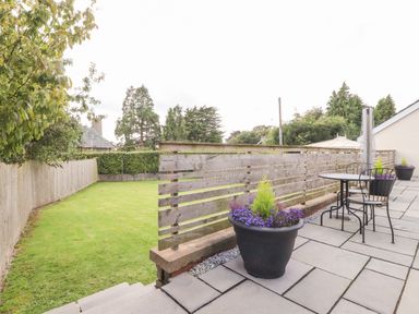 An outdoor patio area with a table and chairs large flower pots and a wooden fence overlooking a grass lawn at Underfell in Burton-In-Kendal