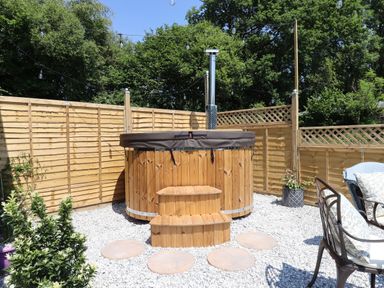 An outdoor area with a wooden hot tub surrounded by a wooden fence and garden chairs at Two Moors Shepherds Hut in Hittisleigh