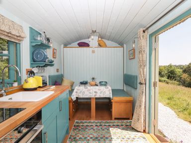 A compact kitchen and dining area inside a small cabin with wood countertops and open doors showing a grassy outside at Two Moors Shepherds Hut in Hittisleigh
