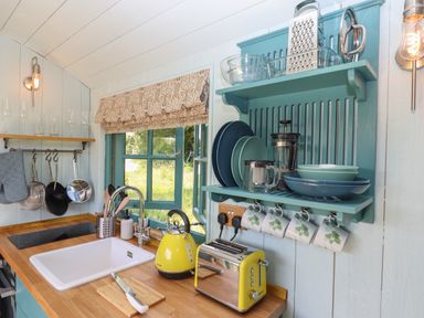 A kitchen with a sink yellow kettle yellow toaster and shelves with dishes at Two Moors Shepherds Hut in Hittisleigh