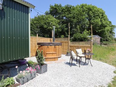 An outdoor area with a green hut a wooden hot tub two chairs and a small table with flowers at Two Moors Shepherds Hut in Hittisleigh