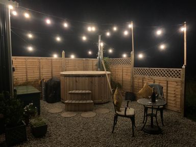 An outdoor patio area with a wooden hot tub string lights and a small table with chairs at Two Moors Shepherds Hut in Hittisleigh