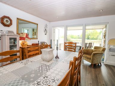 A dining room with a table covered with patterned cloth surrounded by wooden chairs and a cushioned armchair near a glass door at Links View in Fritton