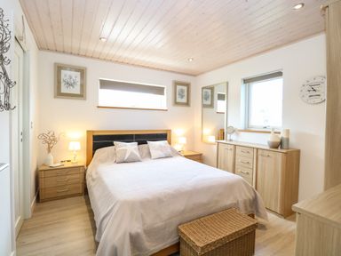 A bedroom with a double bed side tables lamps a chest of drawers mirror and windows at Links View in Fritton