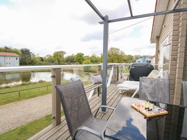 A balcony with chairs, a table with a board game, a barbecue grill, and a bench overlooking a pond and houses at Links View in Fritton