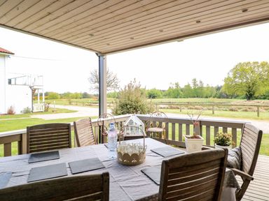 A covered outdoor dining area with wooden chairs around a table set with placemats and a water bottle overlooking a grassy landscape at Links View in Fritton