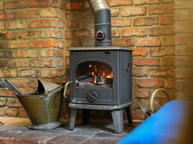 A wood burning stove with fire inside and a metal coal bucket beside it against a brick wall
