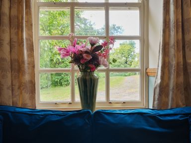 A vase with flowers on a windowsill behind a blue couch with curtains on either side at Manor Cottage in Northallerton