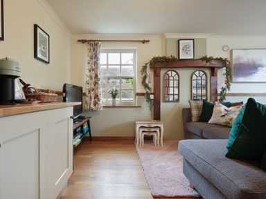 A living room with a gray sofa and green cushions a wooden cabinet a small TV two nested white tables a window with floral curtains and a flower vase at Manor Cottage in Northallerton