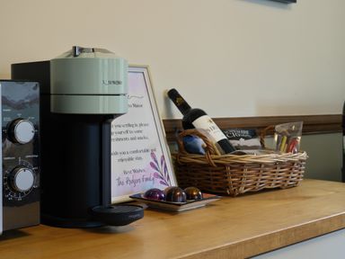 A coffee machine next to a plate with coffee pods a framed welcome note a basket with snacks and a bottle of wine on a wooden counter at Manor Cottage in Northallerton