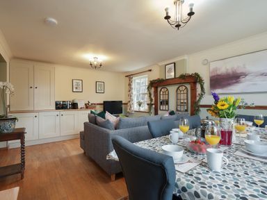 A dining table set with glasses of orange juice bowls and mugs next to a seating area with gray couches in a living room at Manor Cottage in Northallerton