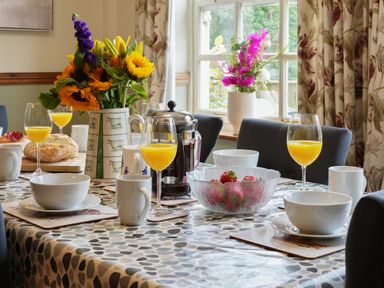 A dining table set for breakfast with bowls cups glasses of orange juice a bowl of strawberries a vase of flowers and a window with floral curtains at Manor Cottage in Northallerton