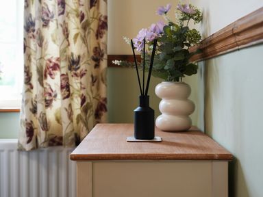 A wooden table with a black diffuser and a white vase with flowers near floral curtains at Manor Cottage in Northallerton